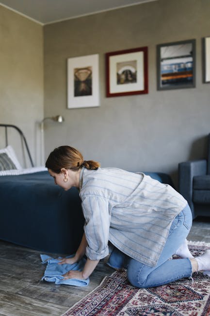 A woman with brown hair tied back, dressed in a light blue striped shirt and jeans, is kneeling on a patterned rug in a living room, cleaning a wooden floor with a blue cloth. The room features a bed 